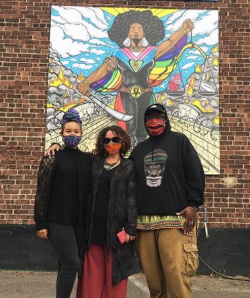 Three people wearing facemasks stand in front of painting mounted on brick wall